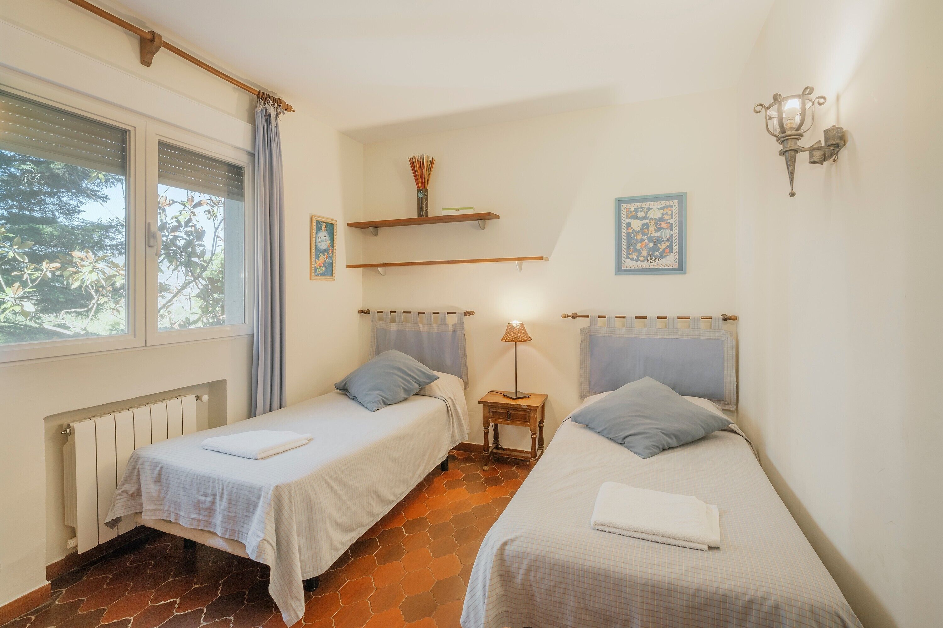 Ein Zimmer mit zwei Einzelbetten, einem Heizkörper, einem Fenster mit Vorhängen, einem kleinen Tisch mit Lampe und Regalen an der Wand.