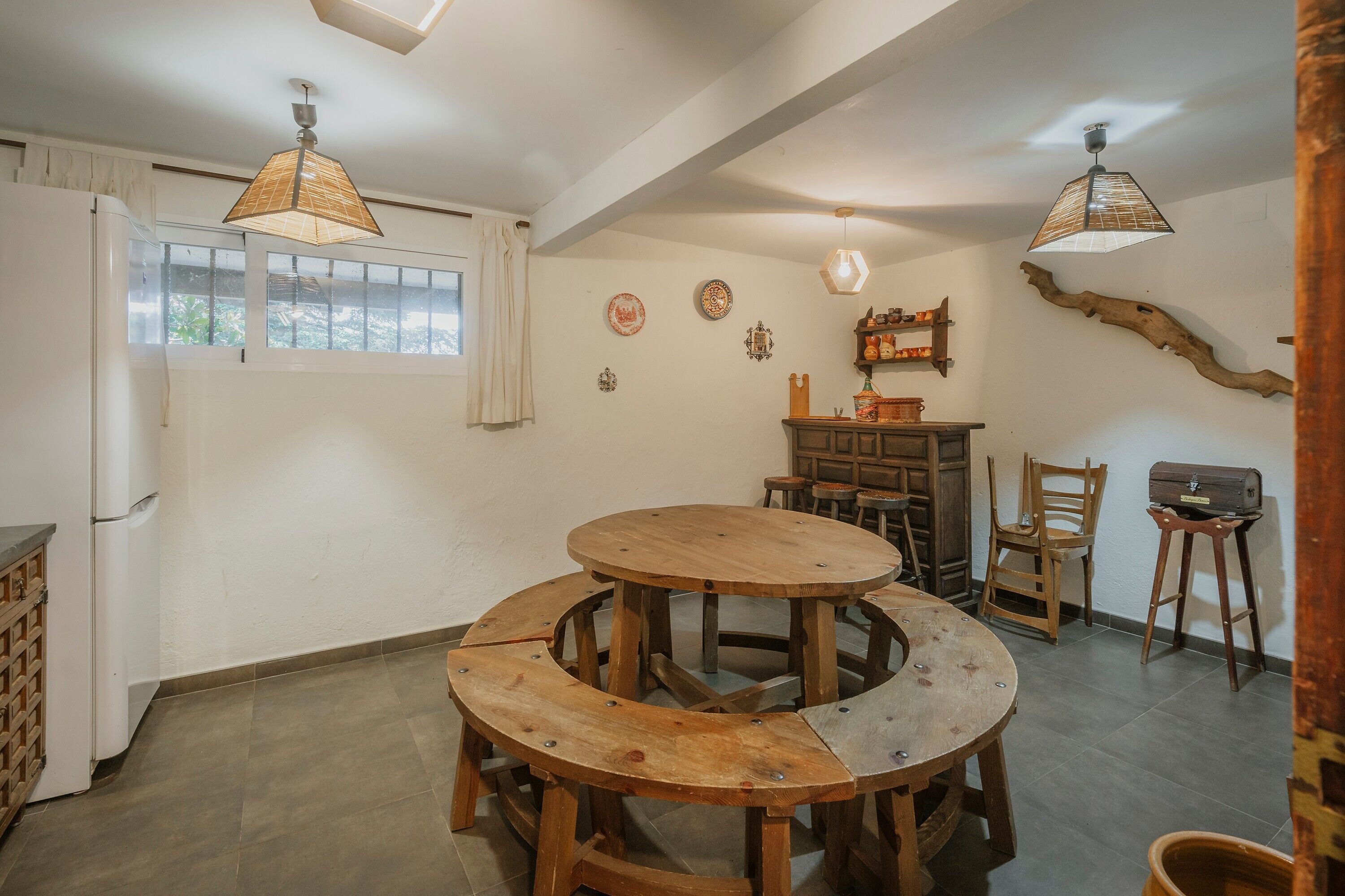 Een keuken met een ronde houten tafel, twee hangende lampen, een koelkast en een raam met gordijnen.