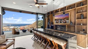 A modern kitchen with a bar area, large windows, and a view of the city and ocean. - Brand-New Villa with Full Staff, 2 Pools, & Close to Downtown Cabo (Cabo San Lucas)