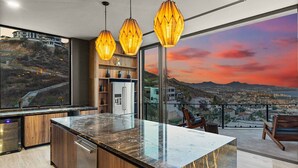 A modern kitchen with a marble island, wooden cabinets, and pendant lights. A large window offers a city view. - Brand-New Villa with Full Staff, 2 Pools, & Close to Downtown Cabo (Cabo San Lucas)