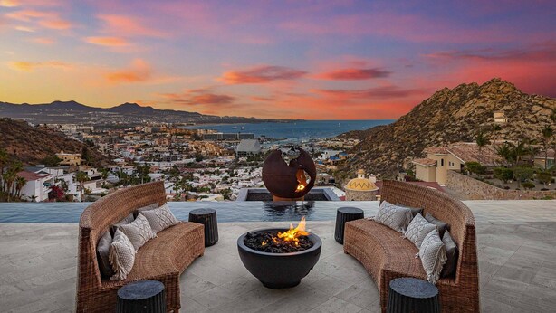 A patio with wicker furniture and a central fire feature, overlooking a coastal cityscape during sunset. - Brand-New Villa with Full Staff, 2 Pools, & Close to Downtown Cabo (Cabo San Lucas)