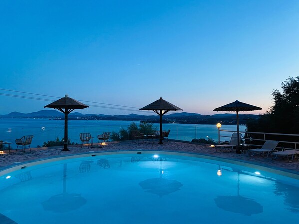 Piscina con sillones de descanso y sombrillas, rodeada por una terraza de piedra, con vista a un cuerpo de agua y montañas distantes.
