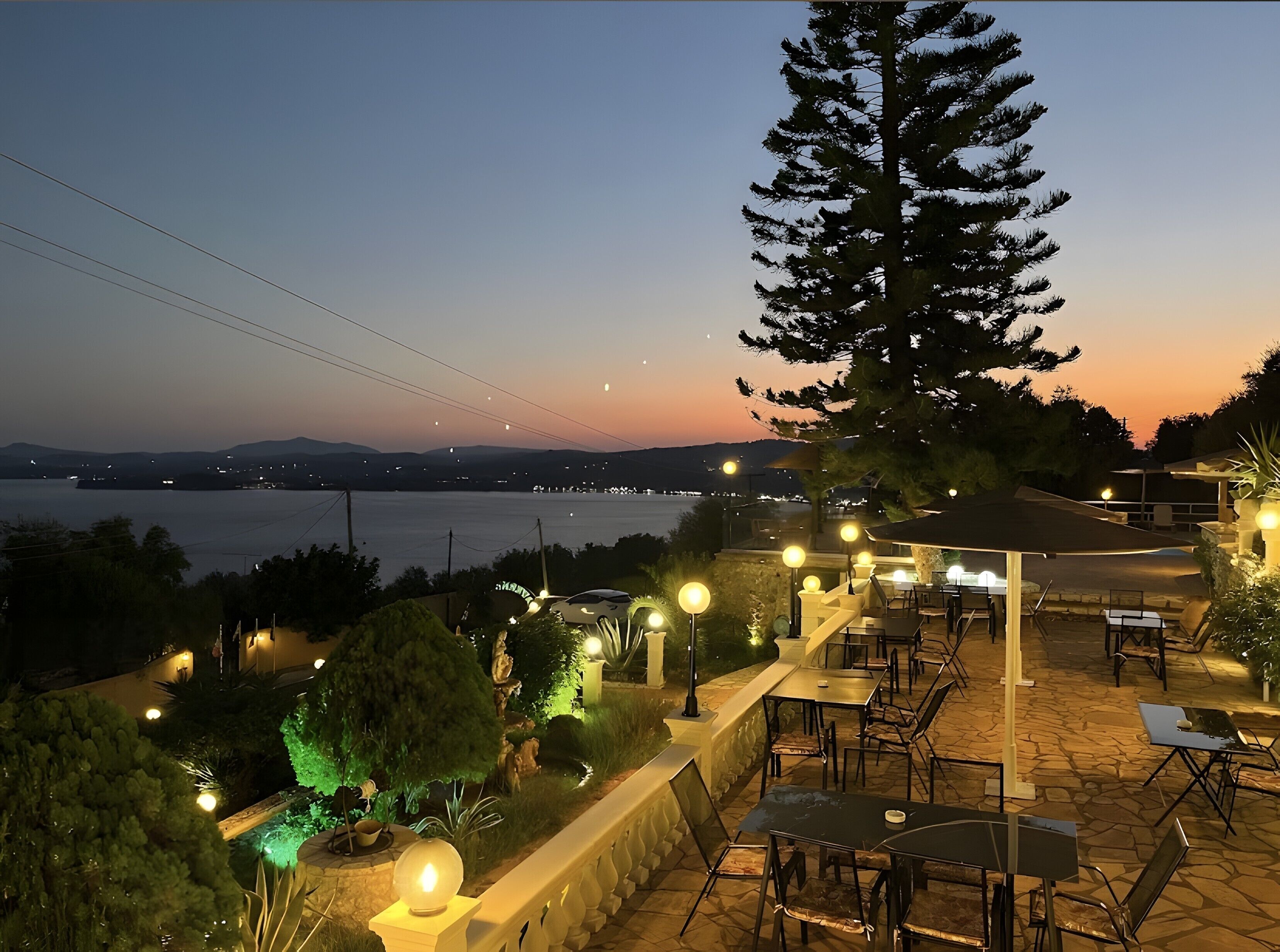Una terrazza esterna con tavoli e sedie, un albero grande e vista sul mare e luci lontane.