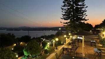 Una terrazza esterna con tavoli e sedie, un albero grande e vista sul mare e luci lontane.
