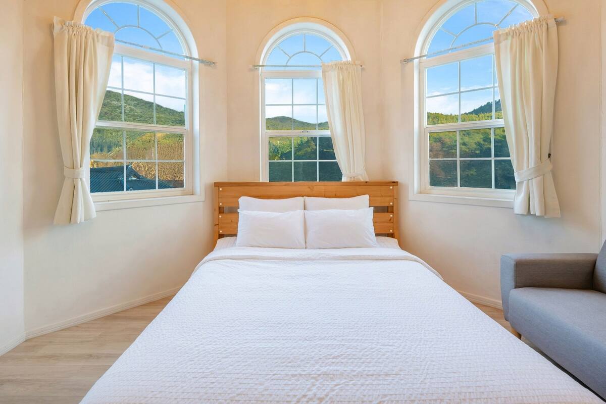 A bedroom with a large bed, a wooden headboard, and three windows with curtains.