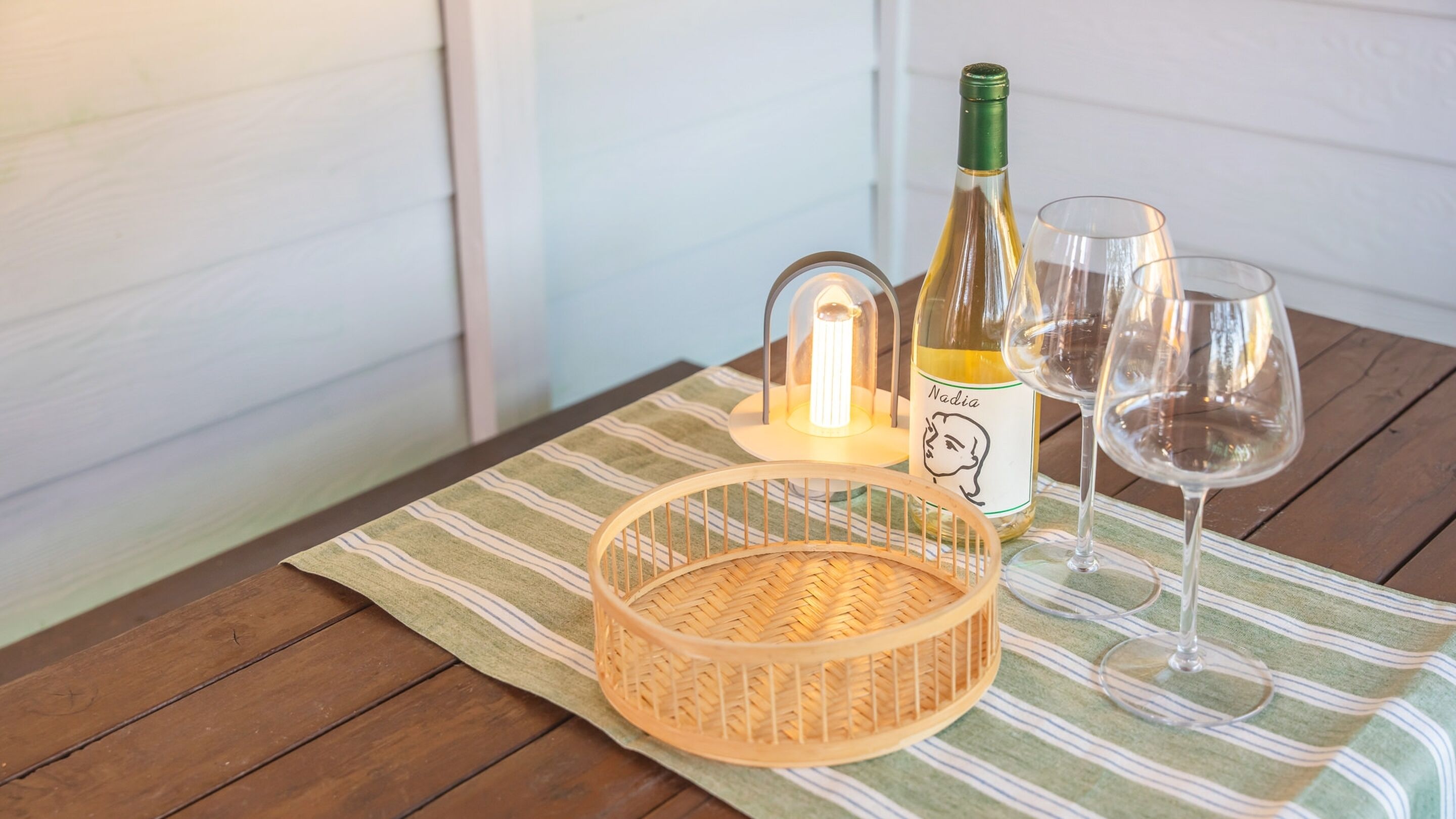 Una mesa de madera con una cesta tejida, una botella de vino, dos copas y un accesorio de iluminación decorativo.