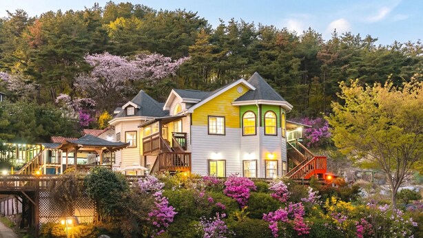 A multi-story house with a prominent yellow and white facade, surrounded by lush gardens and flowering trees. - Gyeongju Woojin Green Pension (Gyeongju)