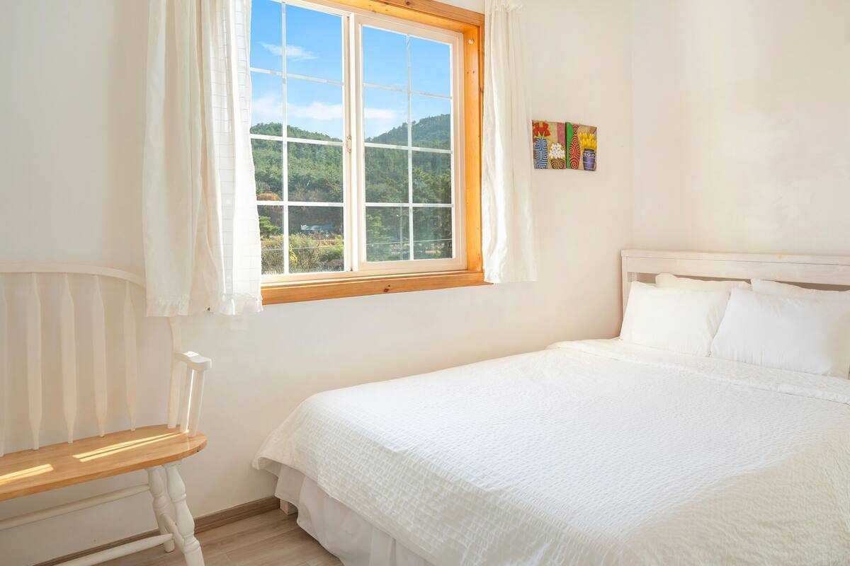 A bedroom with a large bed, a window with a view of greenery, and a small table.