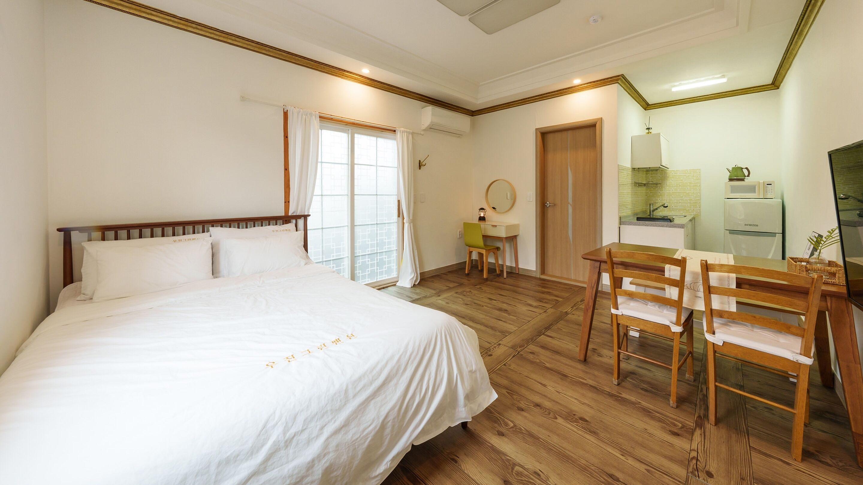 A hotel room with a large bed, a desk, a chair, a small table, a mirror, and a kitchenette.