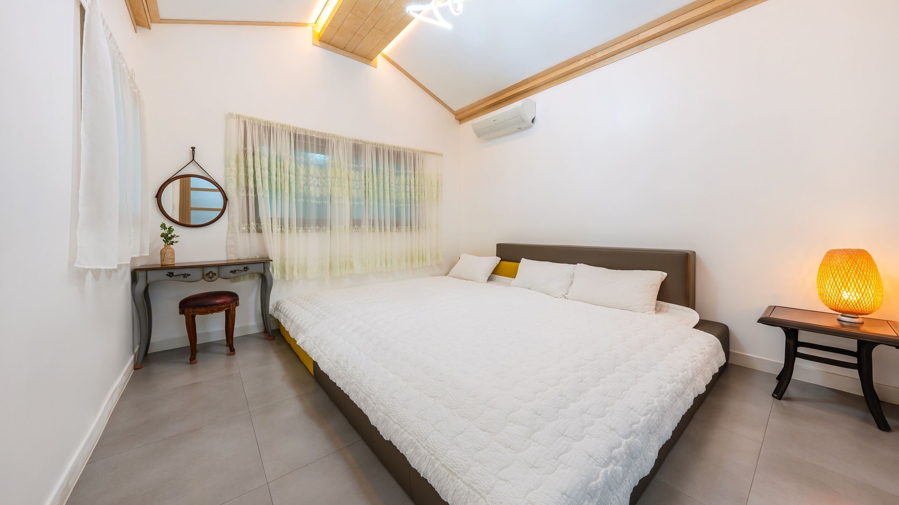 A bedroom with a large bed, a bedside table with a lamp, a dresser, a mirror, and a window with sheer curtains.