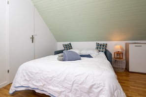 A bedroom with a sloped ceiling, a bed with a white duvet, patterned pillows, a small bedside table with a lamp, and a wooden nightstand.