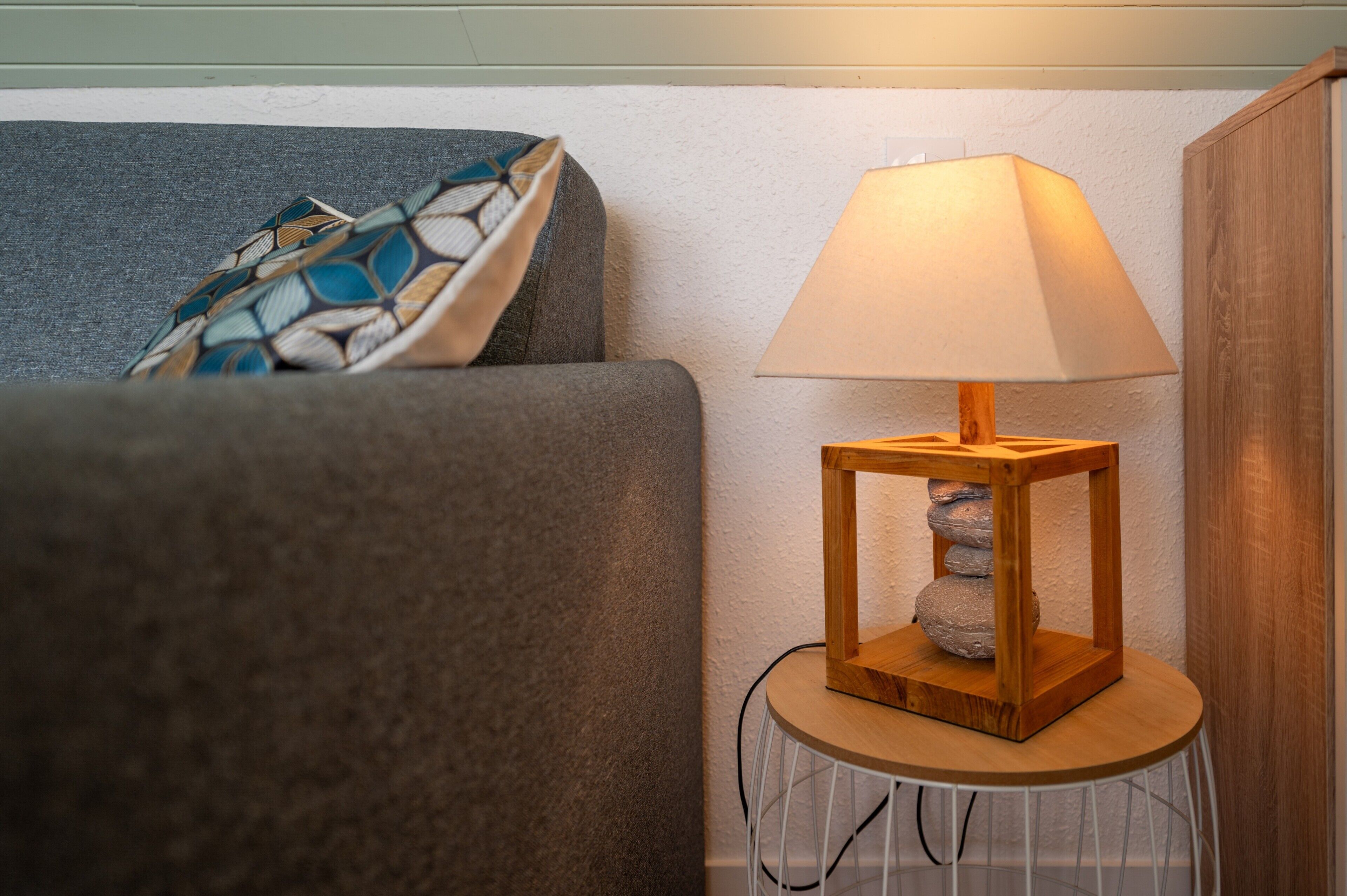 A room with a grey sofa, a patterned throw pillow, a wooden lamp on a small table, and a white cable.