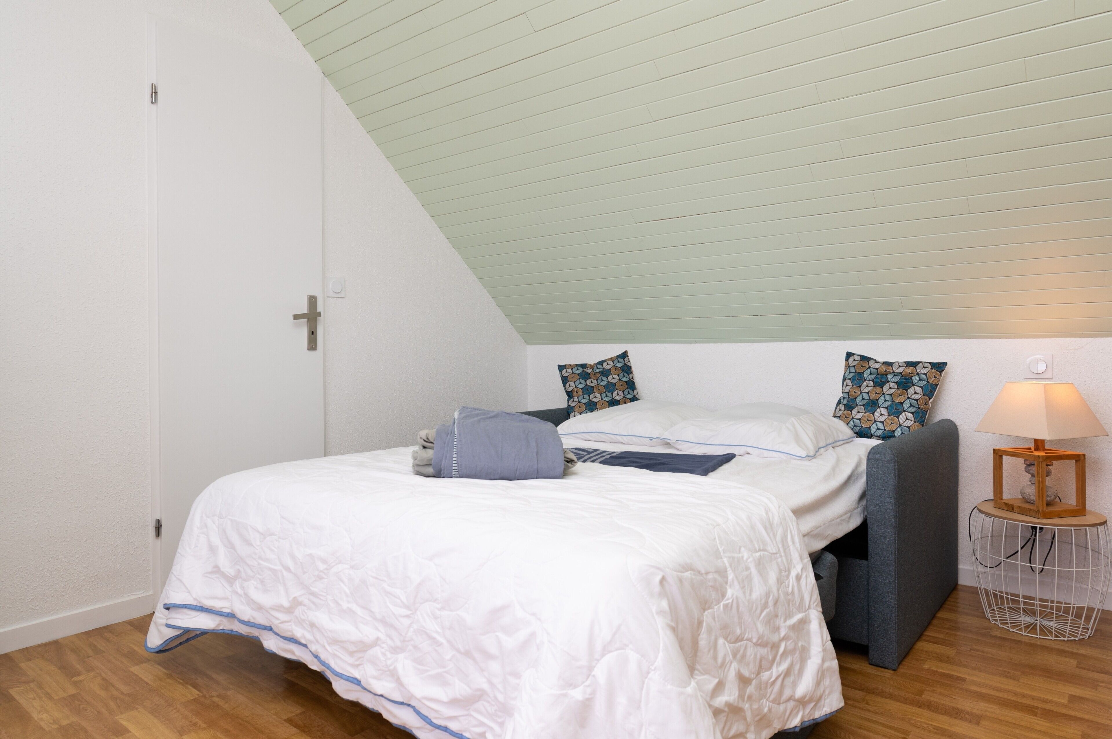 Un dormitorio con techo inclinado, una cama con edredón blanco y almohadas azules, un banco tapizado gris y una lámpara de noche sobre una base de alambre.