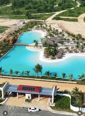 An aerial view of a resort with a large central pool, palm trees, and a small beach area.