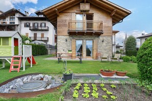 Une maison à deux étages avec un balcon, un espace de jeux doté d’un toboggan et un jardin avec des plantes.
