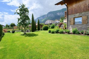 A well-maintained garden with a variety of plants and trees, a stone building with wooden shutters, and a mountainous backdrop.