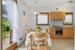 A dining area with a table set for four, a kitchen with wooden cabinets, and a view of a garden through a window. - Chalet 'Agritur La Crucola -' with Mountain View, Wi-Fi and Air Conditioning (Flavon)