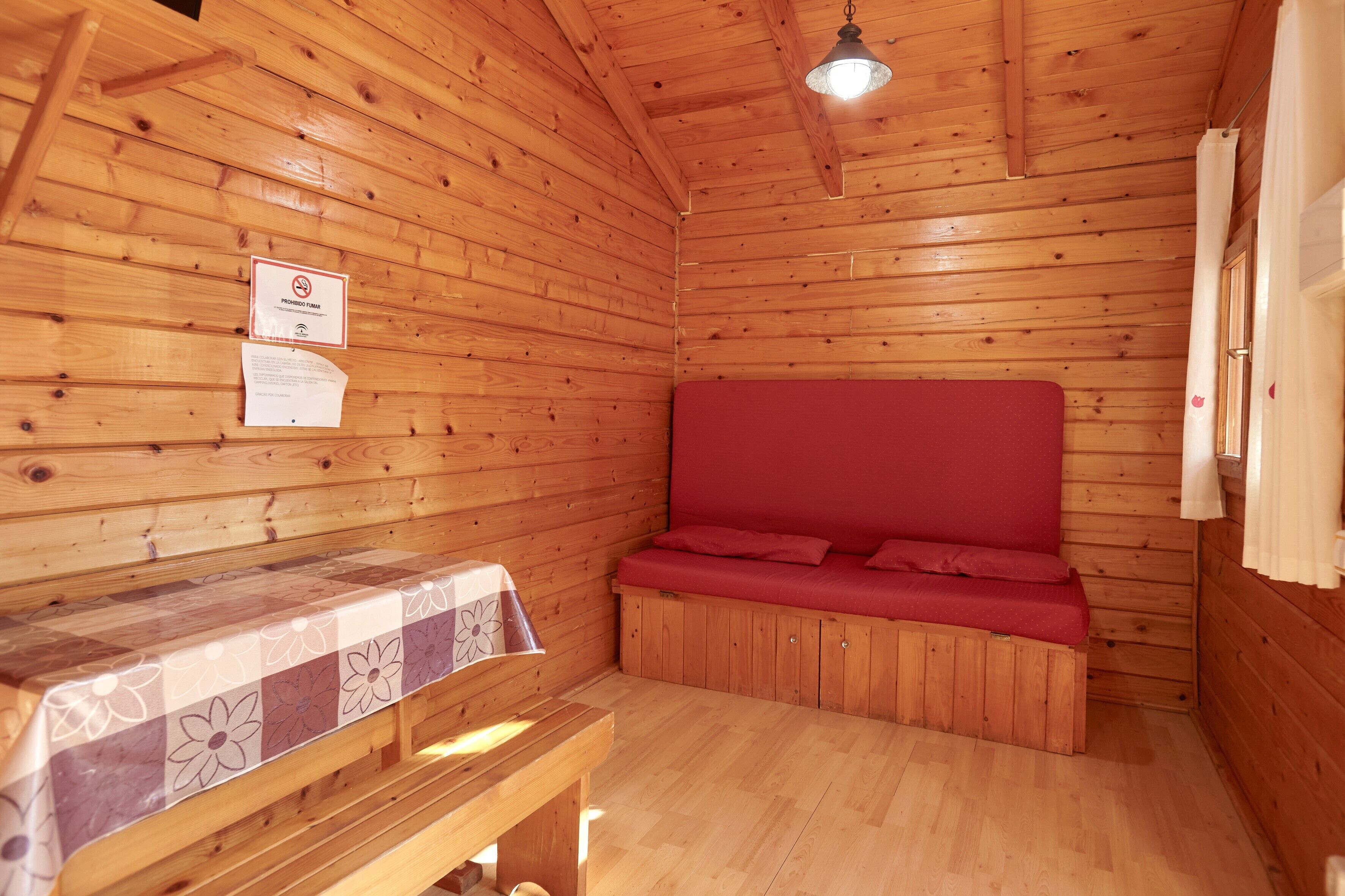 Interior de una cabaña de madera con un sofá rojo, un banco de madera y una mesa con mantel floral.