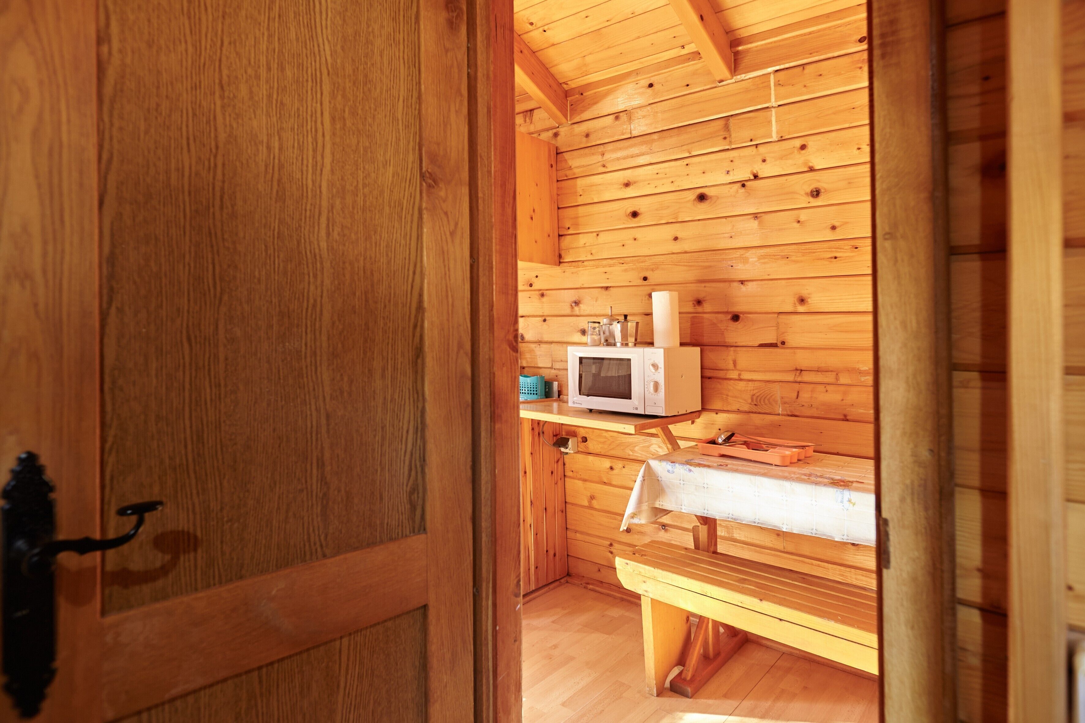 Interior de una cabaña de madera con una puerta de madera, un microondas en un estante y un banco.