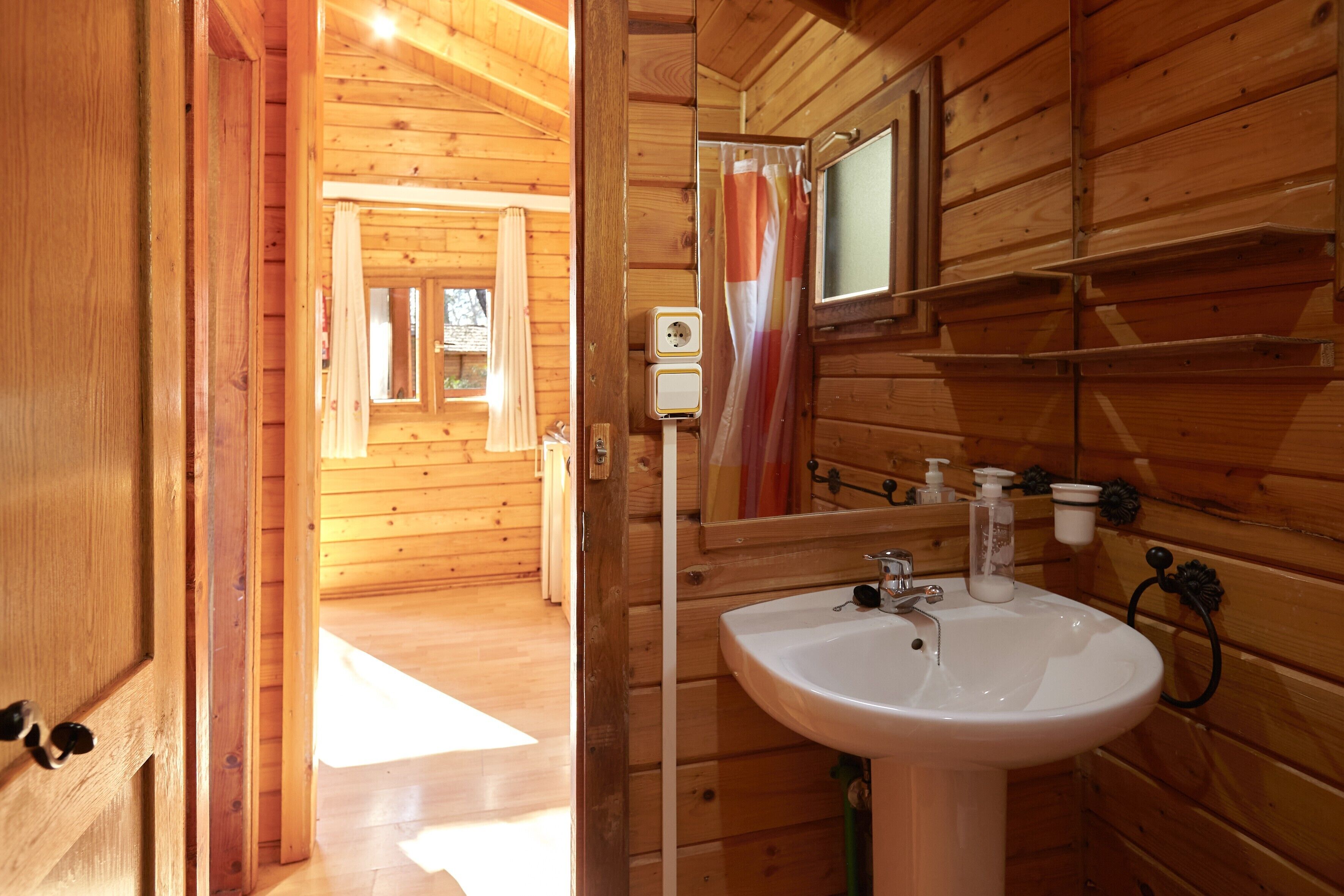 A wooden cabin interior with a wooden door, a white sink, a mirror, and a window with curtains.