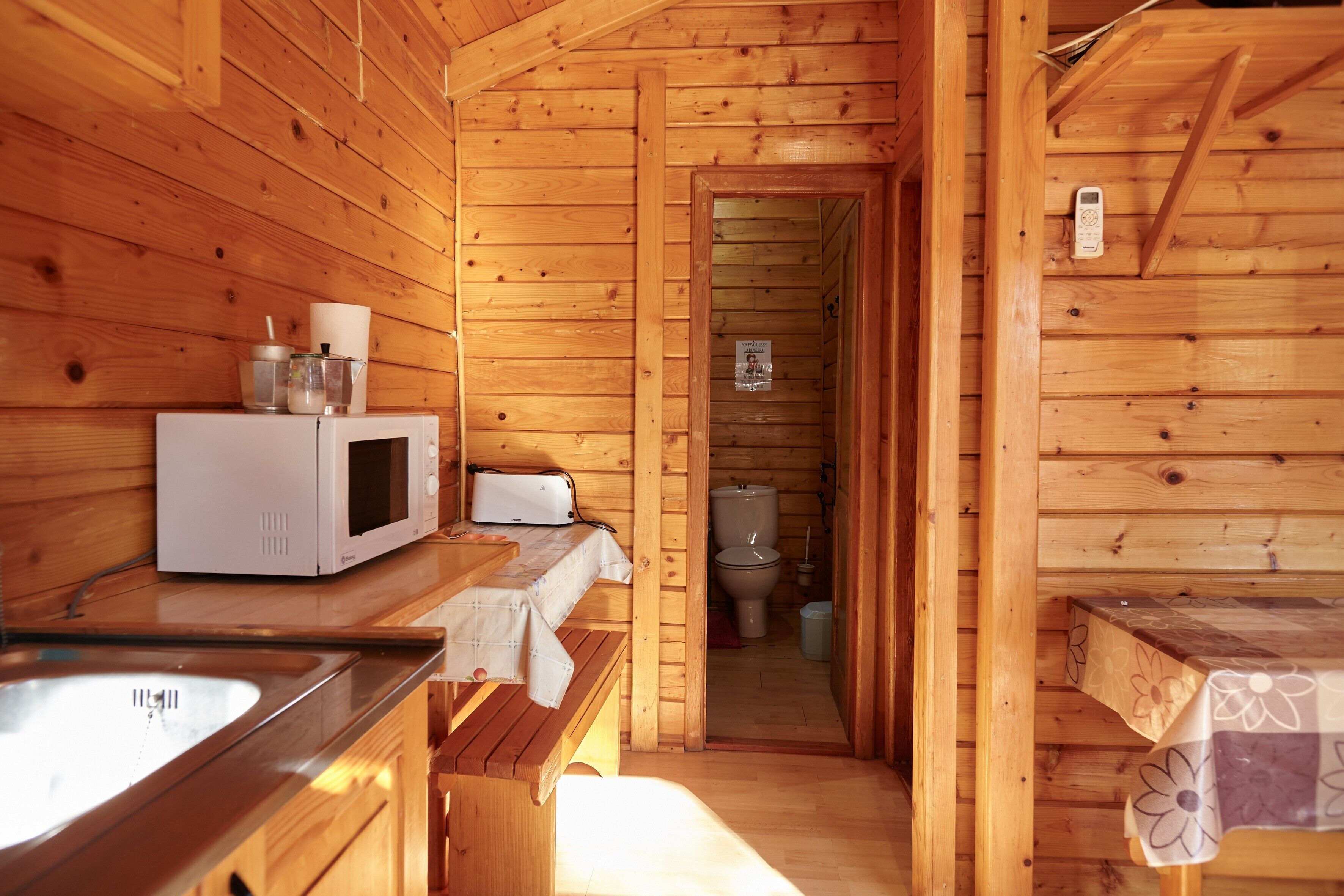 Interior de una cabaña de madera con cocina, microondas, lavamanos y un inodoro visible a través de un vano abierto.