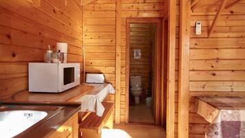 Interior de una cabaña de madera con cocina, microondas, lavamanos y un inodoro visible a través de un vano abierto.