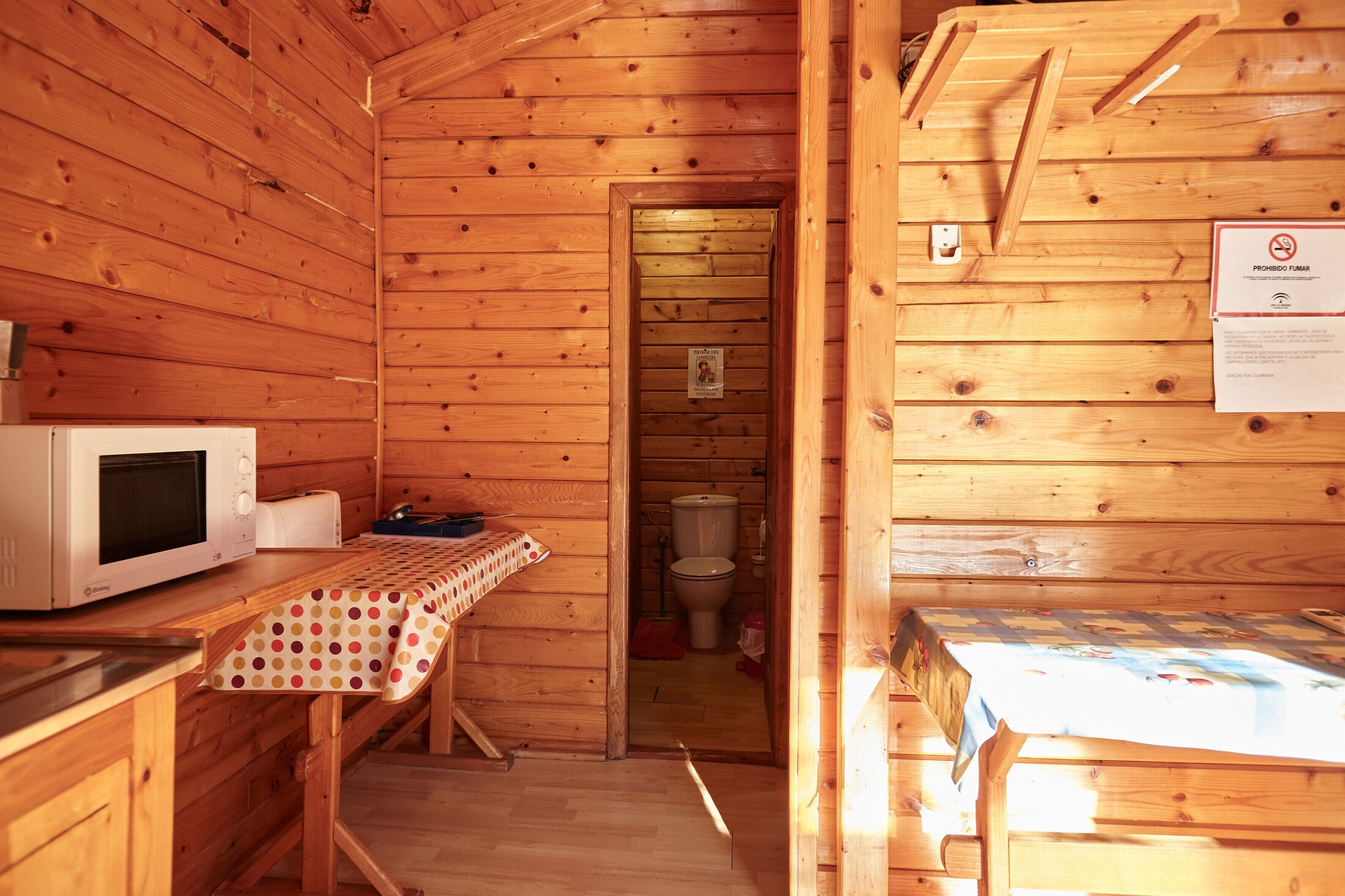 A wooden cabin interior with a kitchen area, a dining table, and a bathroom visible through an open door.
