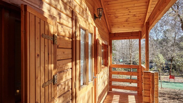 Une cabane en bois dotée d’une véranda, de grandes fenêtres et offrant une vue sur la forêt environnante.