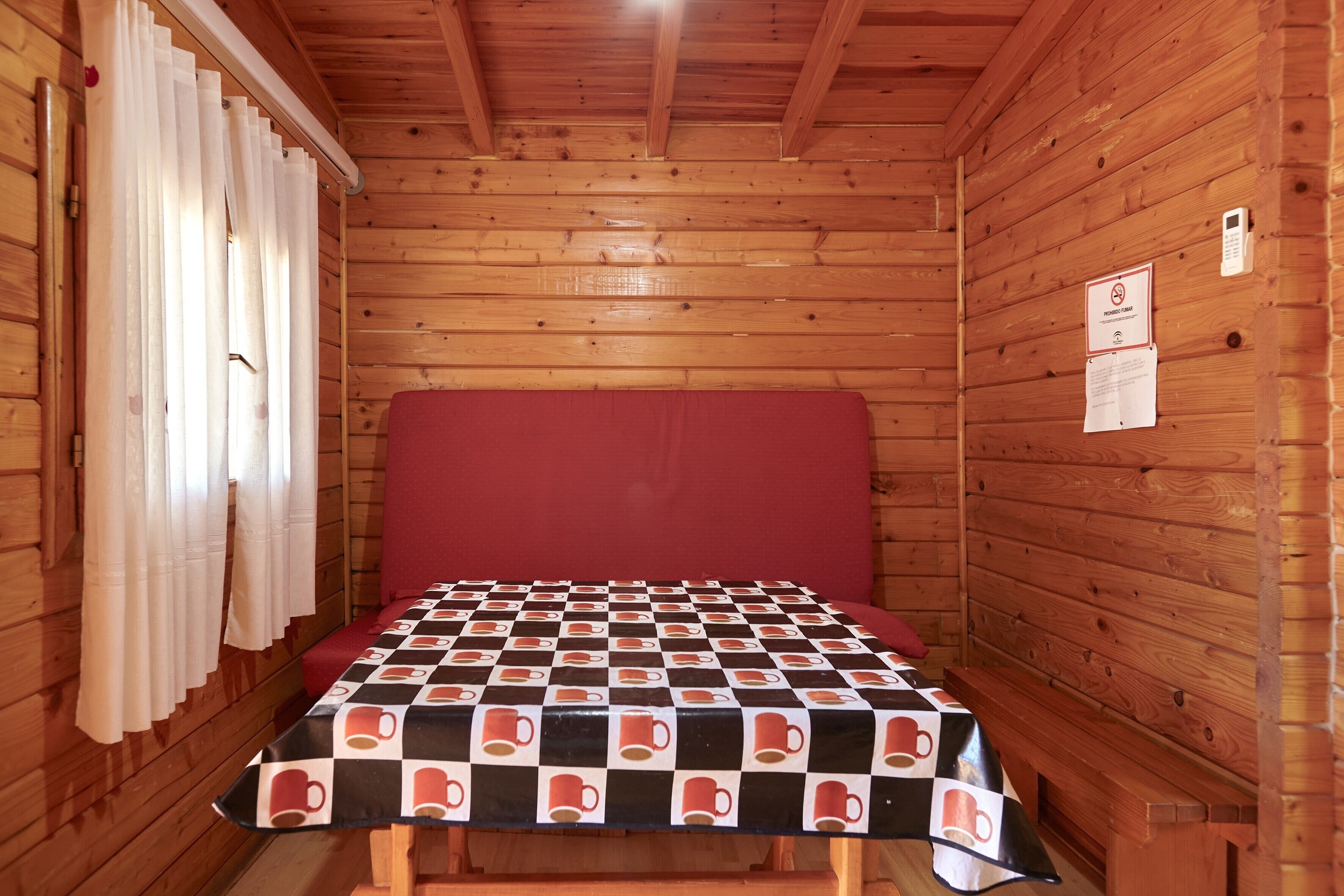 A small wooden cabin room with a checkered tablecloth, a red cushioned bench, and a wooden ceiling.