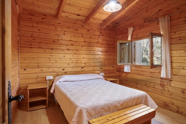 Un dormitorio de cabaña de madera con cama, un banco, una ventana con cortinas y una lámpara en el techo.