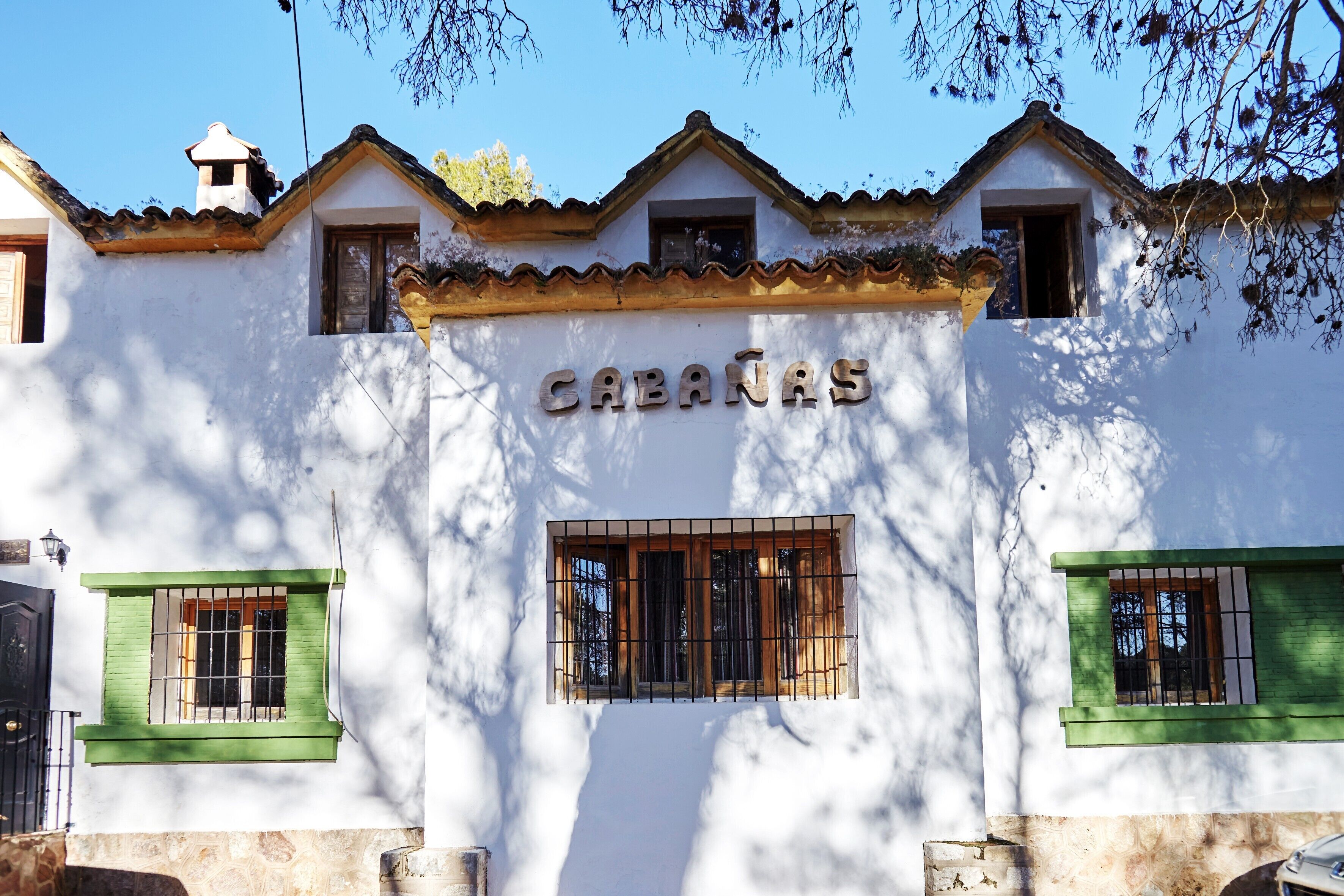 Un edificio con el nombre 'CABANAS' en la fachada, con dos ventanas con persianas verdes, un techo de tejas y una pequeña torre con campanario.