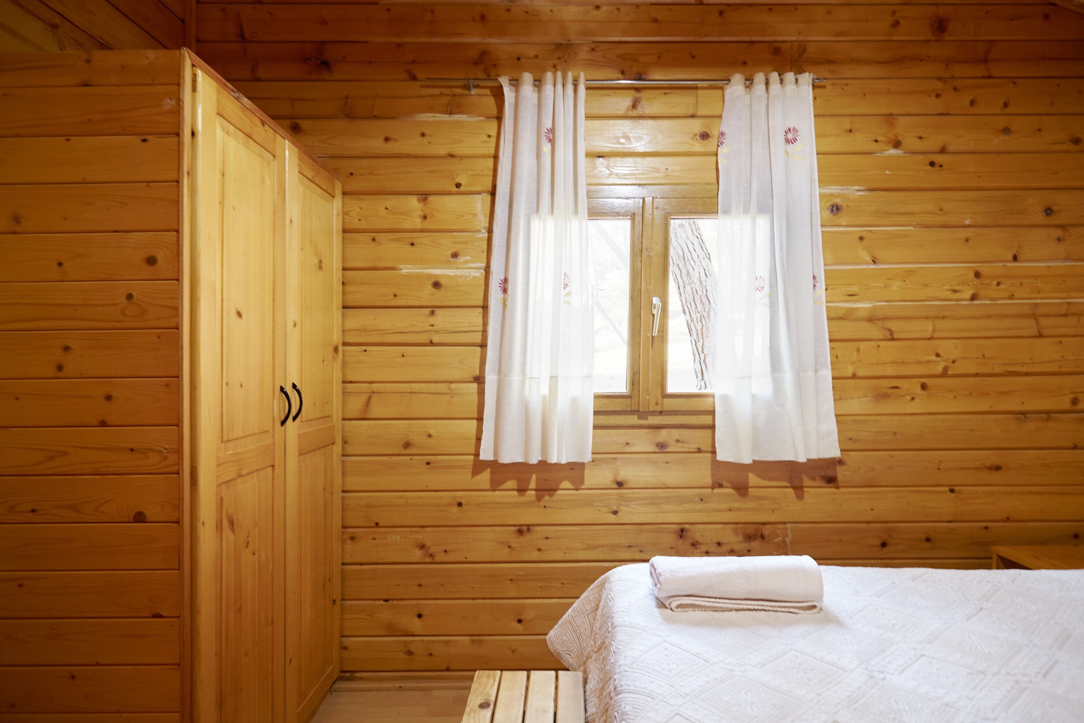 Una habitación de cabaña de madera con una cama, un armario y dos ventanas con cortinas.