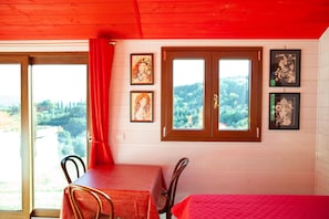 A room with a wooden ceiling, a dining table with chairs, a bed with a red bedspread, and two windows with curtains.