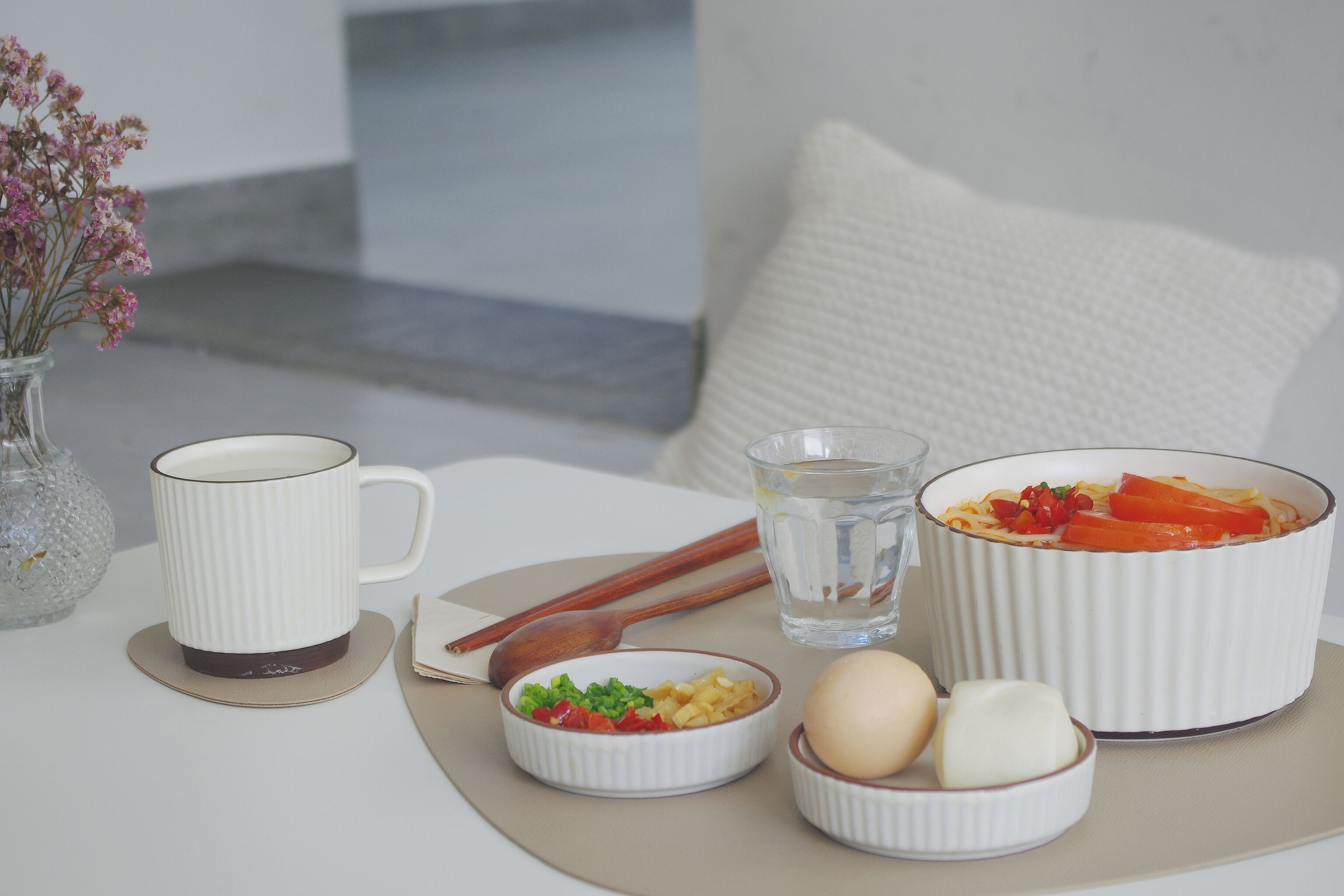 Una mesa con una taza blanca, un plato de comida, un vaso de agua y un jarrón de flores.