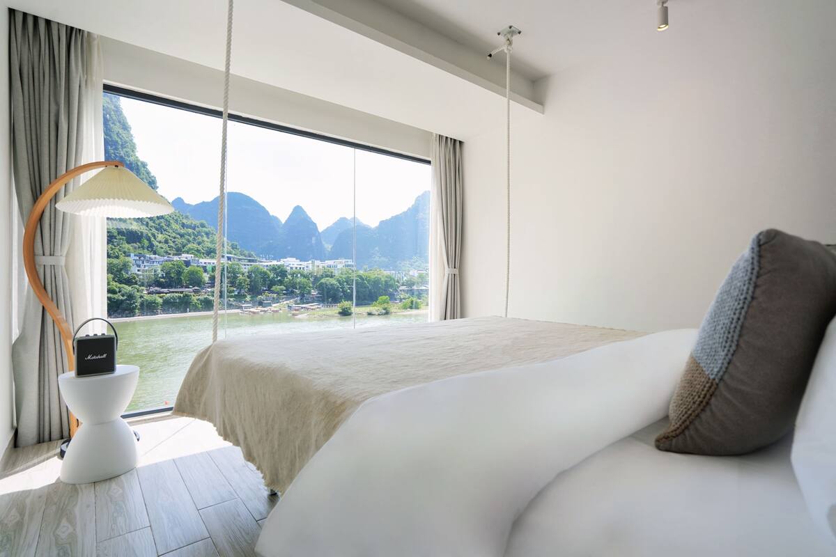 A bedroom with a large bed, a view of mountains and a body of water, a lamp, and a small table.