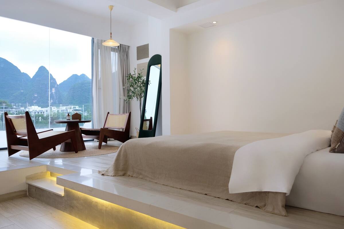 A modern hotel room with a large bed, a seating area with chairs, a dining table, and a view of mountains.