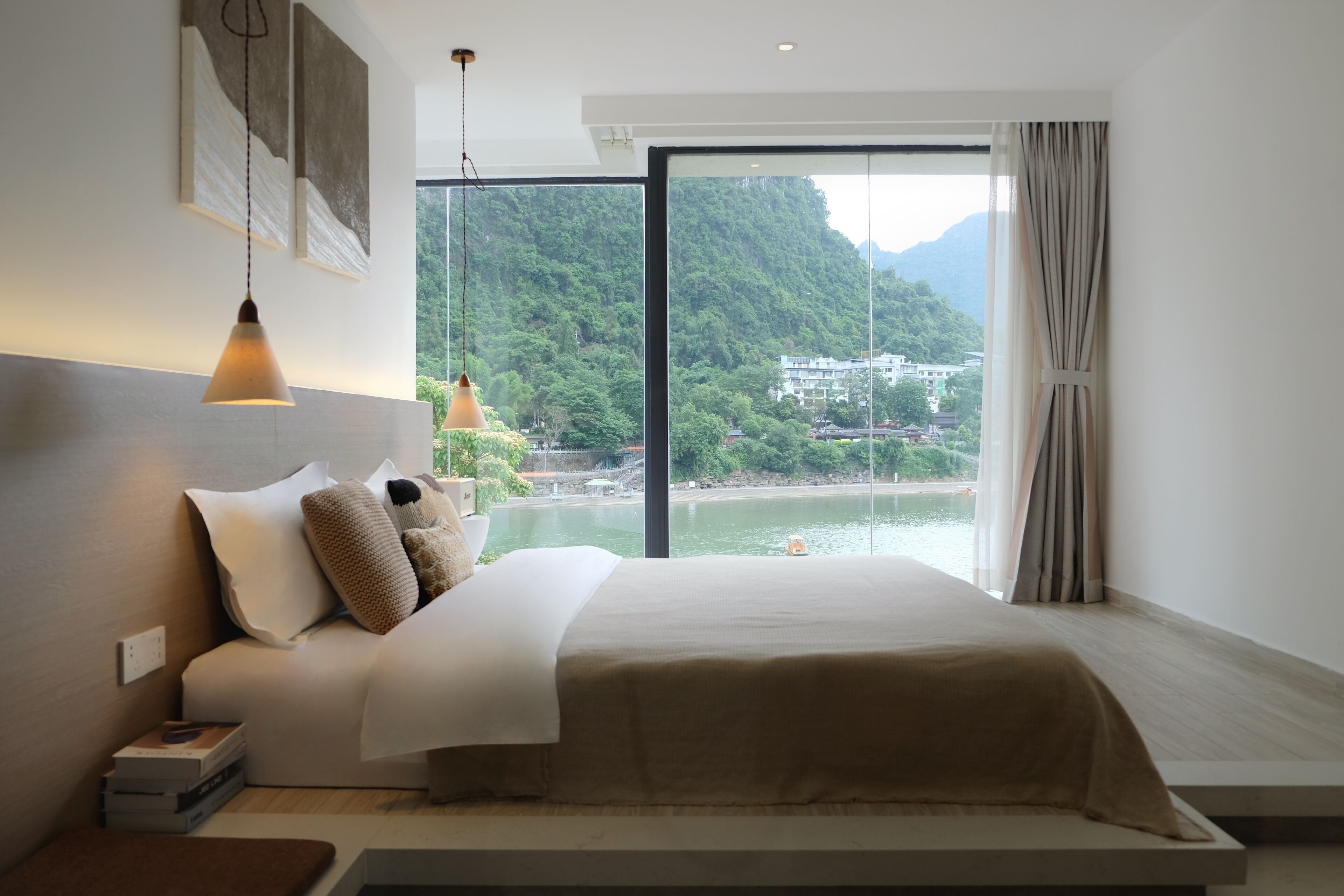 Habitación de hotel moderna con una cama grande, una mesita de noche con libros y vistas a un lago y montañas.