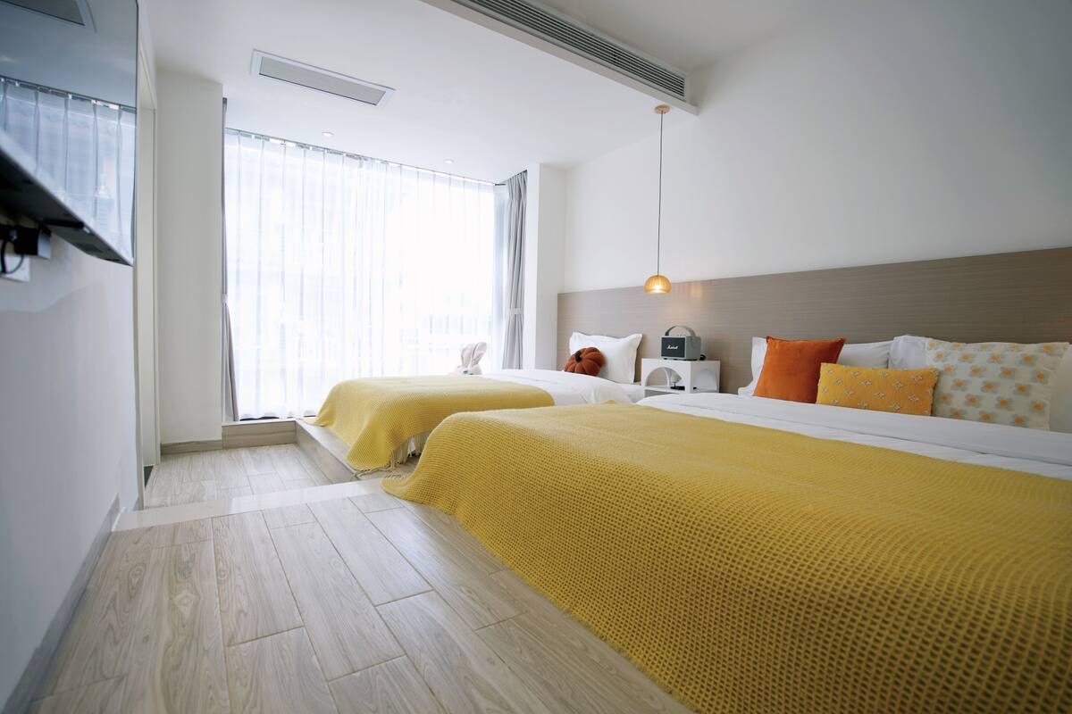 A hotel room with two beds, a large window with curtains, a bedside table with a lamp, and a headboard with a light fixture.