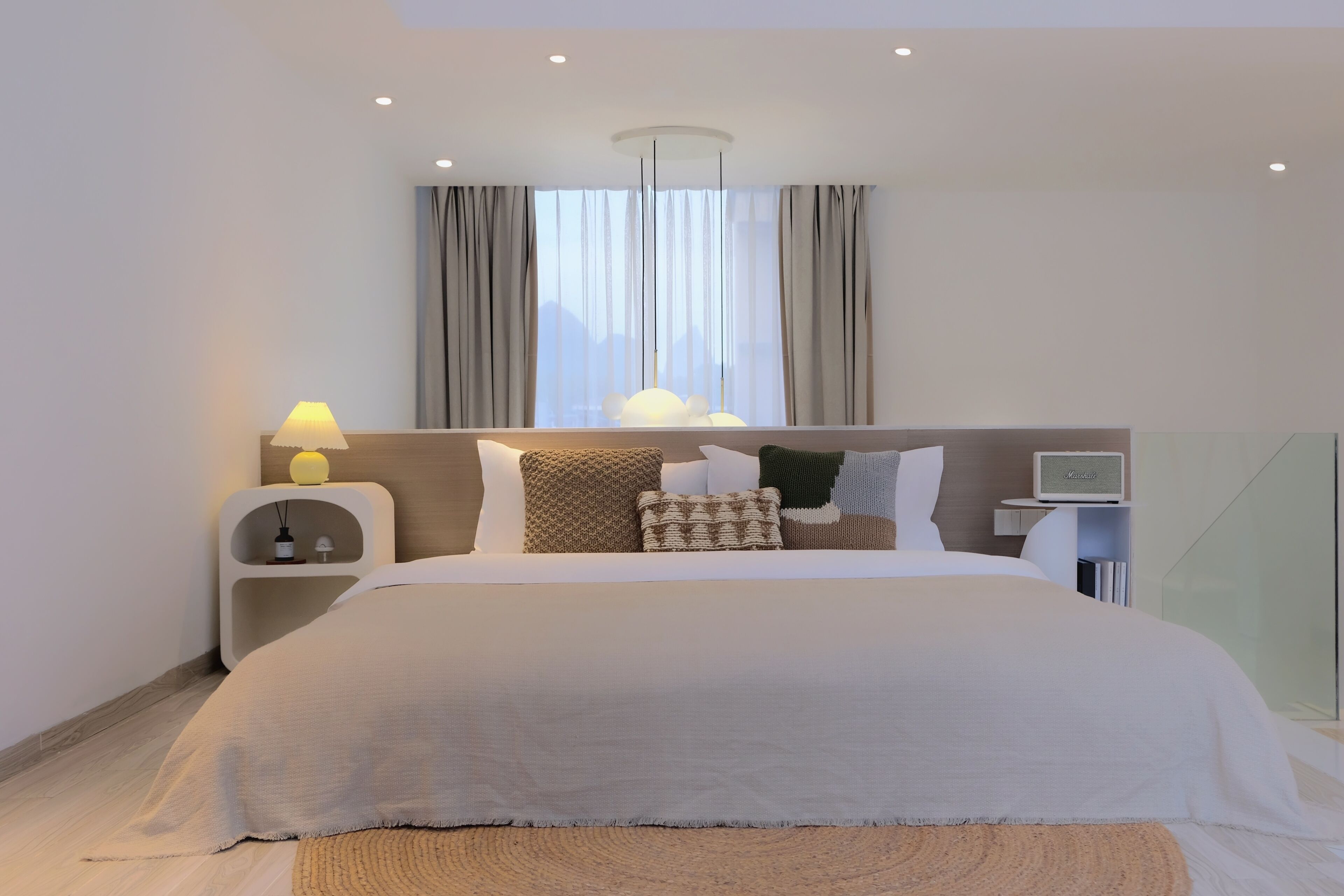 Un dormitorio moderno de hotel con una cama grande, una mesita de noche con lámpara, un estante pequeño y una ventana con cortinas.
