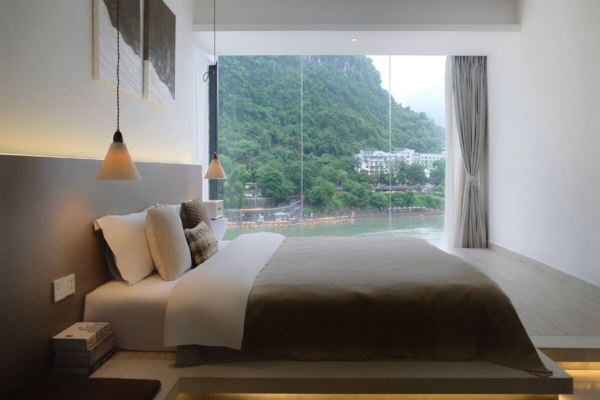 A modern bedroom with a large bed, a view of a river and mountains, and a hanging light fixture.