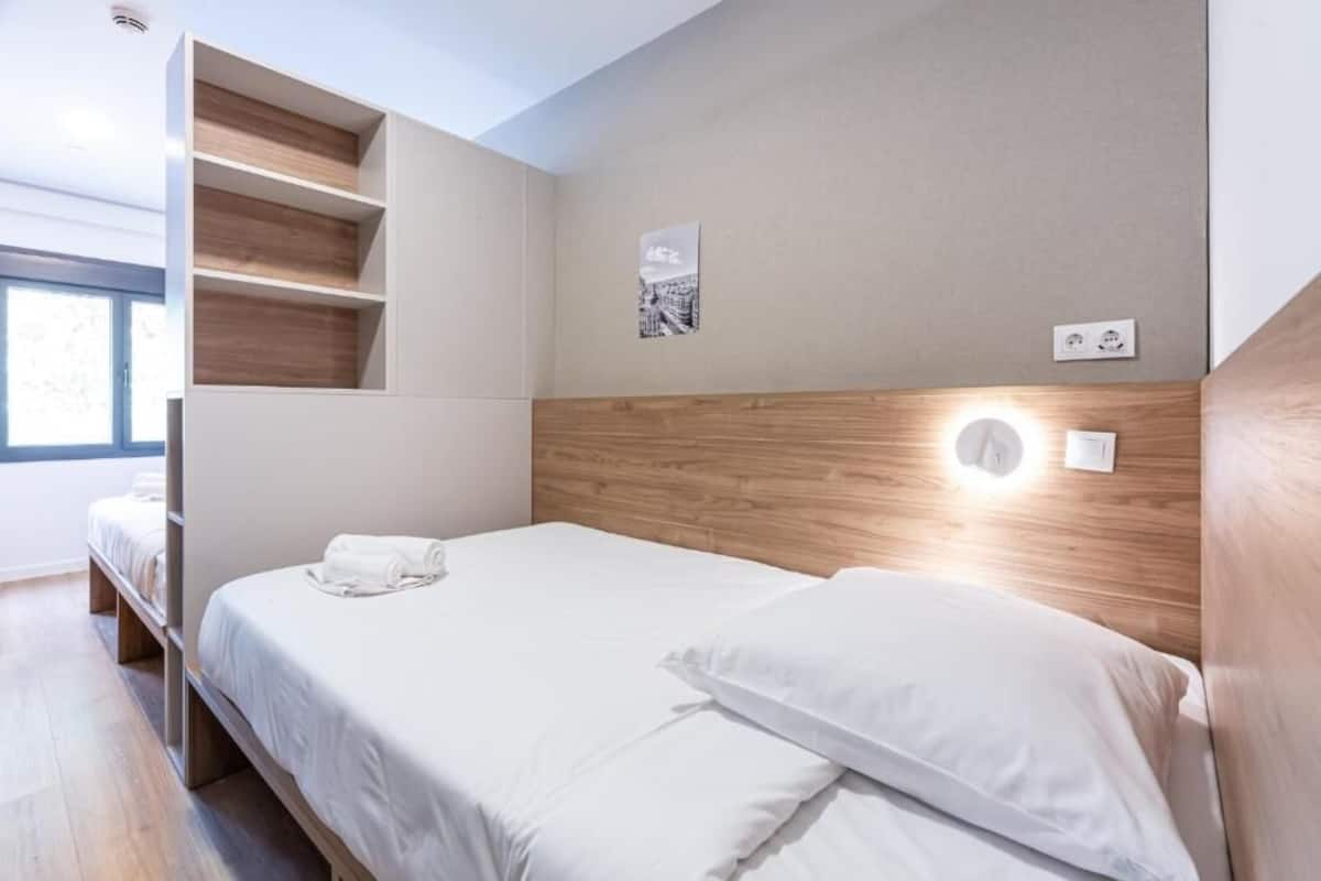 Une chambre d’hôtel moderne, équipée d’un lit, d’étagères intégrées, d’une table de chevet, d’une lampe fixée au mur et d’une fenêtre.