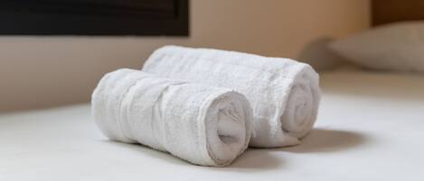 Two rolled white towels on a bed with a plain white headboard and a window in the background.