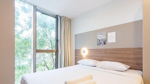 Hotellihuone, jossa on suuri ikkuna, valkoisilla lakanoiden päällä varustettu sänky, puinen pääty ja seinään kiinnitetty valaisin.