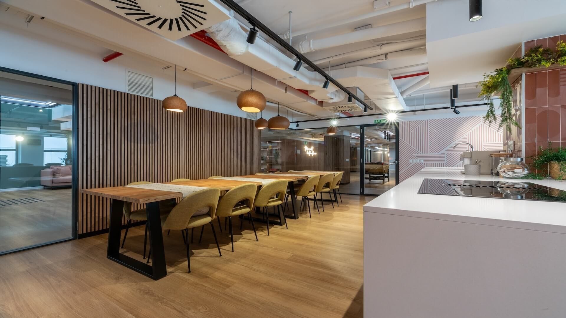 A modern office space with a long wooden table, multiple chairs, pendant lighting, and a kitchen area with a sink and countertop.