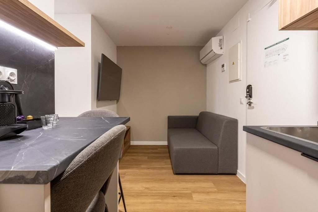 A compact kitchen with a marble countertop, a built-in stove, a grey sofa, and a wooden floor.