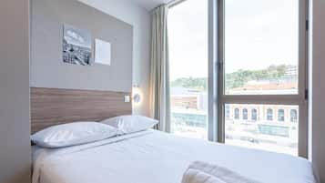 Habitación de hotel con una cama grande, cabecera, ventana con vista a la ciudad y dos cuadros en la pared.