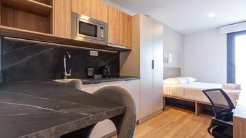 Cocina compacta con microondas, cafetera y un lavamanos pequeño. Hay una cama con colchón y almohadas blancas, y una silla con asiento negro.