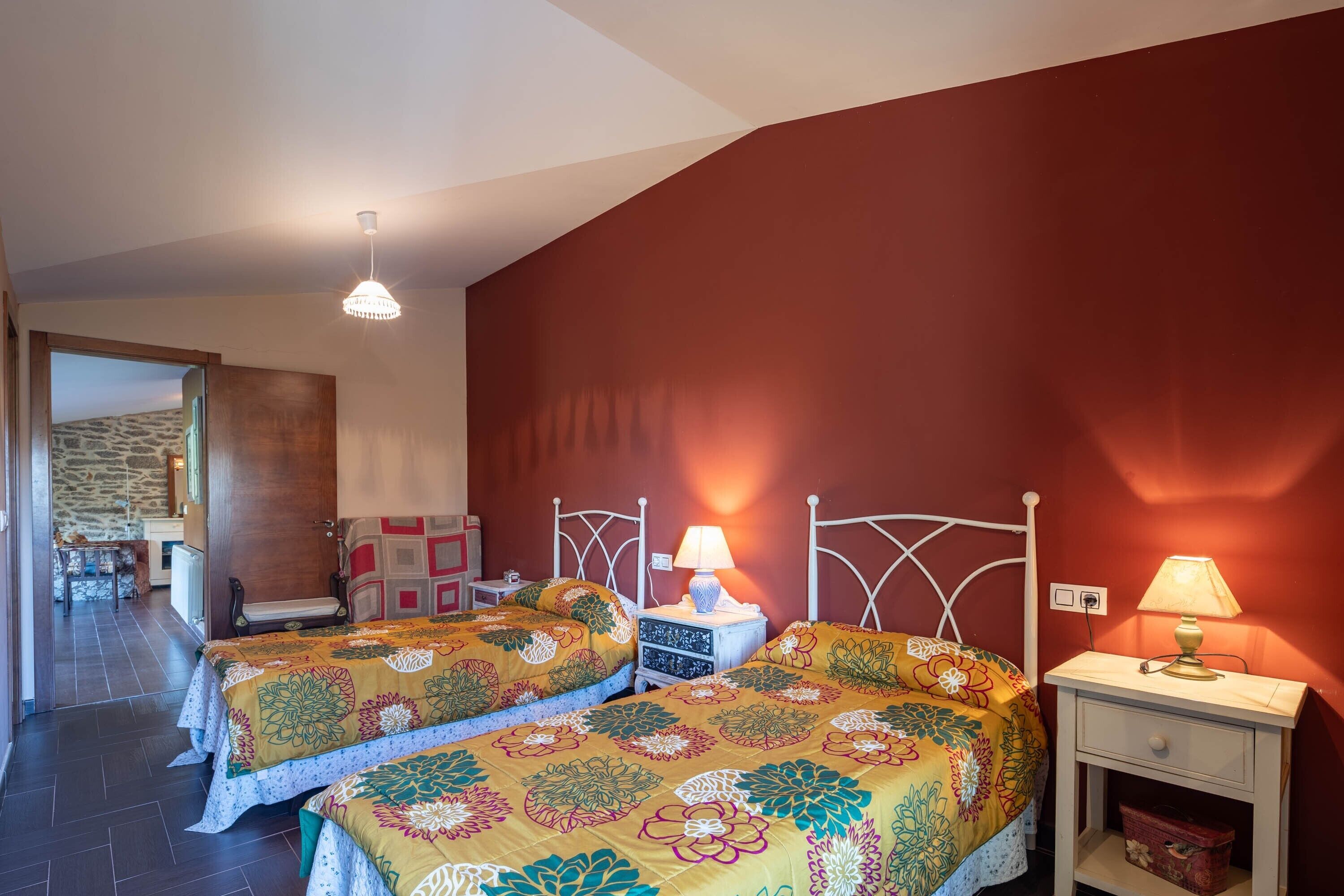 A bedroom with two beds, a nightstand with a lamp, and a view of a stone wall and a table outside.
