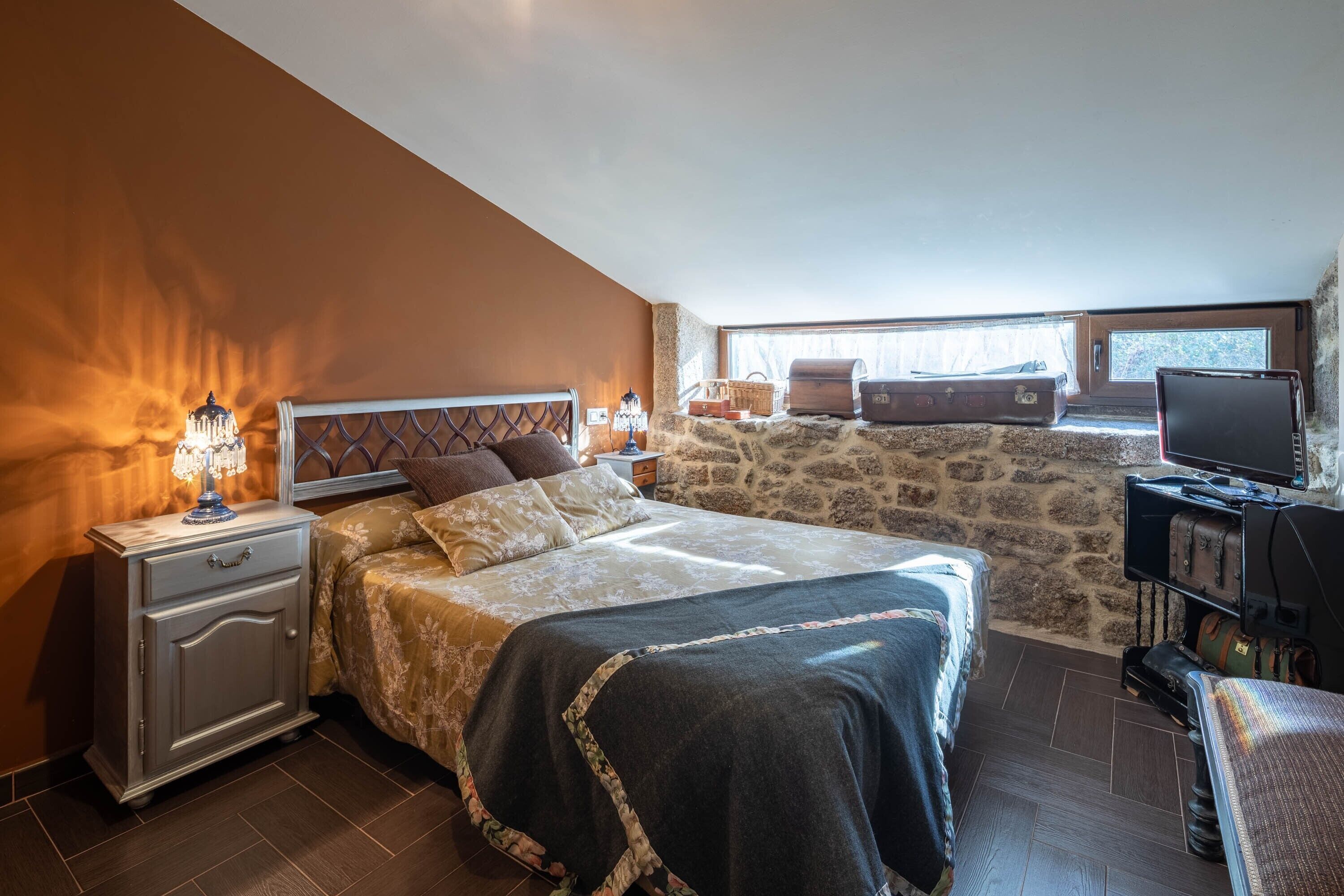 Ein Schlafzimmer mit Steinsturz, einem Bett mit gemustertem Überwurf, einem Nachttisch mit Lampe, einem Schreibtisch mit Computer und einem Fenster mit Blick.