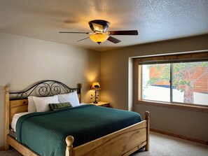 Une chambre à coucher avec un lit en bois, un ventilateur de plafond, une table de chevet avec une lampe et une fenêtre donnant sur des arbres.